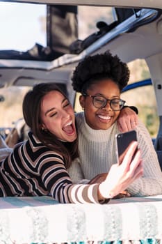 two young women taking a selfie photo lying in a camper van, concept of weekend getaway with best friend and road trip adventure