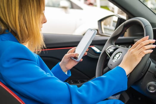 Young smiling woman in the blue suit sitting in the car and texting on her mobile phone