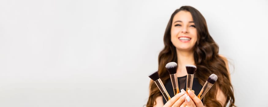 A smiling brunette woman holding makeup brushes on the white background on the banner.