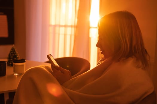 A girl wrapped in a blanket uses a mobile phone in a romantic atmosphere in an apartment with a cozy sunset light from the window, side view