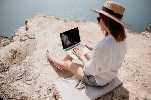 Successful business woman in yellow hat working on laptop by the sea. Pretty lady typing on computer at summer day outdoors. Freelance, travel and holidays concept.