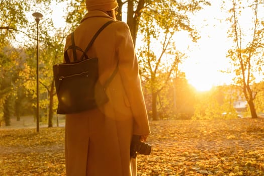 Back view of the woman in a brown coat with a backpack holding a camera against sunlight in the autumn park