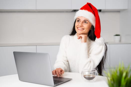 Exciting happy woman in Santa Claus hat imagines happy Christmas or New Year holidays. A woman works at home using her notebook before the party.