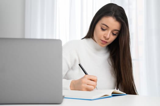 Young woman takes online courses via notebook making notes while listening to teacher. Attentive student sits at table in room studying online at home
