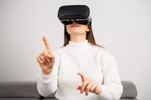 Happy woman in VR glasses moves hand sitting on comfortable couch in apartment living room. Young housewife enjoys modern technology at home