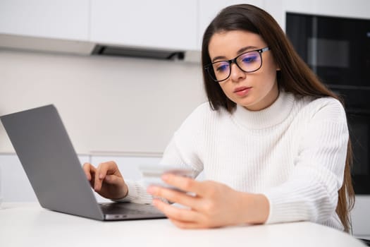 Satisfied woman in glasses enters credit card details onto website via Notebook. Young female customer makes online payment sitting at table in kitchen