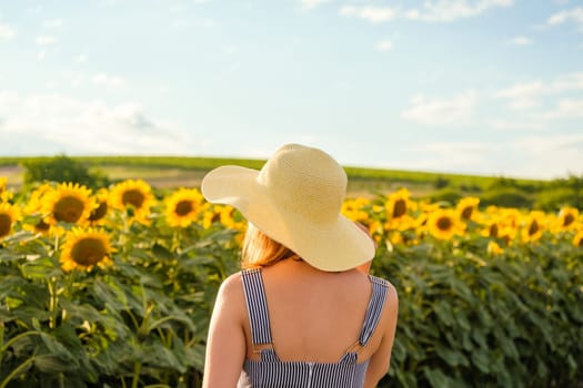 Young woman in wicker straw hat stands against sunflower field on sunny summer day. Lady enjoys nature in countryside backside close view