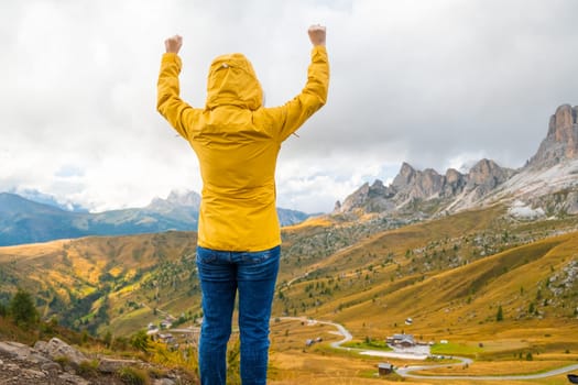 A young woman in the hood and yellow jacket enjoying the mountains view of the Italian Dolomites and raise your hands. Travel concept