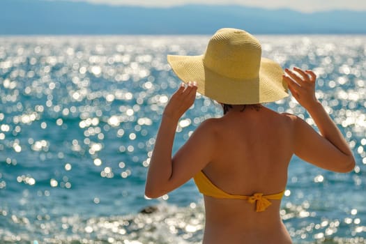Woman in yellow bikini and straw hat looks at blue sea on warm day. Lady holds headdress with hands against water sparkling in sun close back view