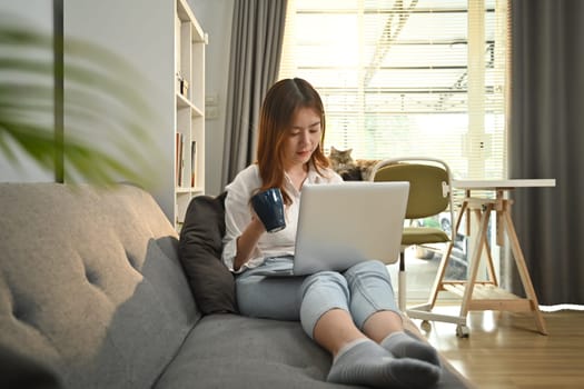 Happy woman relaxing on couch browsing wireless internet or chatting online on laptop computer.