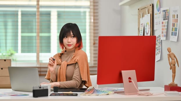 Portrait of young asian woman designer sitting at graphic studio in front of computer monitor and looking confidently to camera.