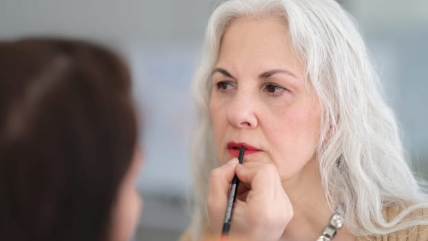 Cosmetologist painting lips with brush to elderly woman with gray hair in beauty salon. Professional makeup services concept