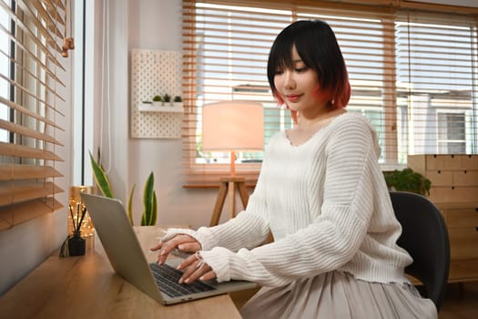 Pretty young Asian woman using laptop browsing internet and checking videos on social networks, sitting in a cozy living room.