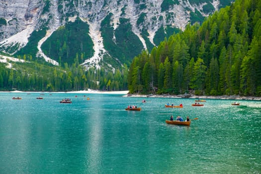 Tourists ride wooden boats on Braies lake with transparent water and Dolomites on the background in sunny day.