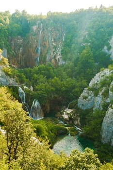 Waterfalls with cascades falling from steep rocks into canyon. Forestry and bare cliffs surround gorge with clear blue lake on bottom of Plitvice lakes upper view