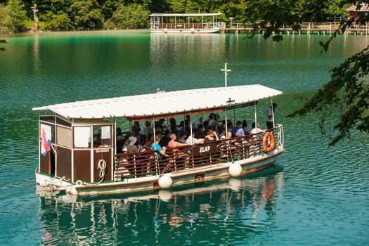 Excursion boat with tourists sails on deep aquamarine lake water. Famous touristic attraction in natural park reserve on sunny summer day on Plitvice lakes