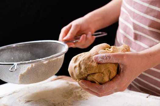 Close up woman sifts wheat flour through a sieve into mixing bowl for dough preparation for cake and bakery on the black background. Knead the dough for homemade pastry.