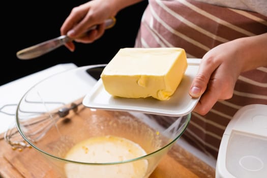 The dough preparation process. Female hands add butter into the mixing bowl for baking cake on a black background. Homemade recipe of pastry.