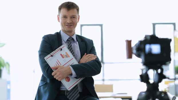 Successful businessman coach with documents with graphs in hands standing near camera in office. Blogging and teleconferencing concept