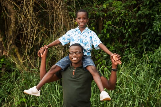 young man in green tee-shirt standing with his child on shoulders smiling on camera.