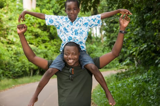 a young man standing in a park with his son on his neck doing gestures while smiling.