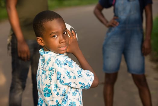 young boy standing with a ball in a park looking sideways smiling and in the background his parents.