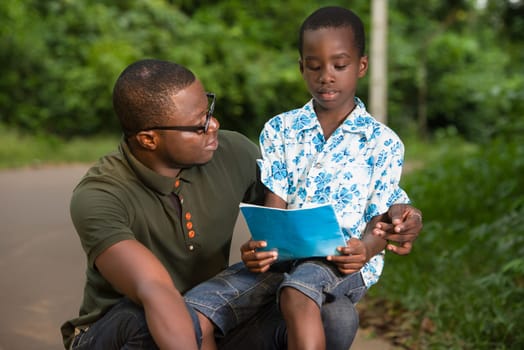a young man crouched in a park watching his child on his knee read.