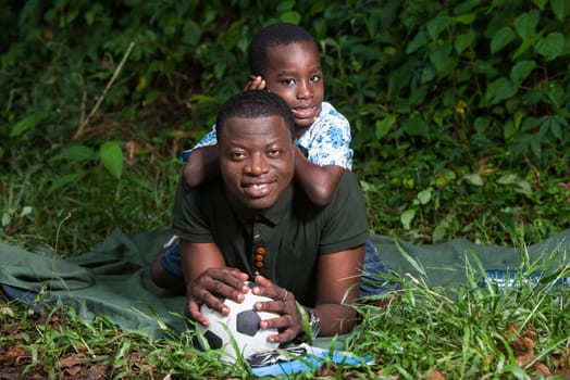 a young man lying in a park with his child on his back watching the camera smiling.
