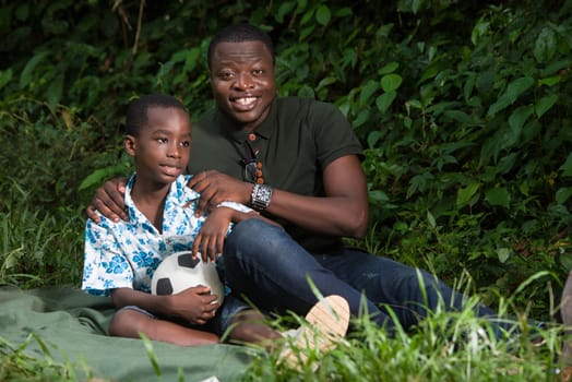 a young man sitting in a park with his son and looking at the camera smiling.