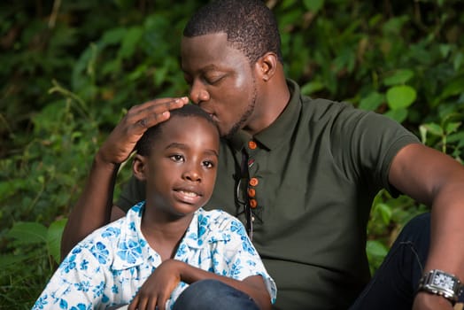 a young man sitting in a park kissing his son's head with his eyes closed.