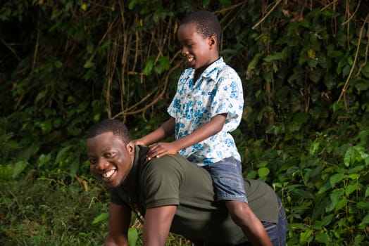 a young man on all fours in a park with his son on his back smiling at the camera.