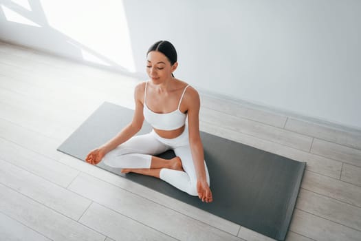 On yoga mat. Young caucasian woman with slim body shape is indoors at daytime.