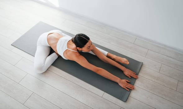 On yoga mat. Young caucasian woman with slim body shape is indoors at daytime.