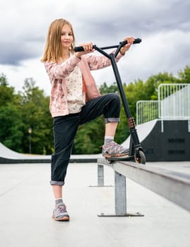 Preteen girl standing with scooter at street in the park and looking at camera. Cute child posing with eco vehicle outdoors