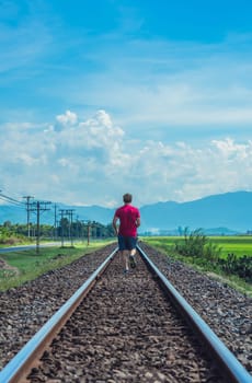 Man runs on railroad tracks bright summer sun day landscape red t-shirt shorts back view.