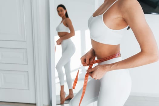 Uses measuring tape. Young caucasian woman with slim body shape is indoors at daytime.