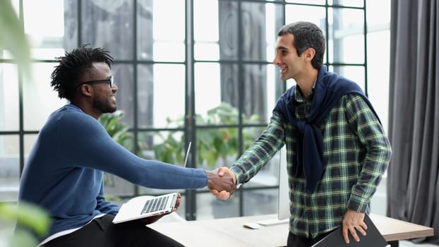 Two smiling businessmen shaking hands together while standing by windows in an office boardroom