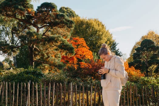 Beautiful young woman takes photos with a professional camera in autumn forest. Smiling girl enjoying autumn weather. Rest, relaxation, lifestyle concept. Young photographer takes pictures of autumn forest at sunset