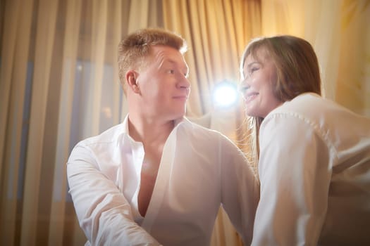 An adult couple of newlyweds in in white shirts in a hotel room after the wedding. A guy and a girl, a man and a woman in beautiful room. The concept of love and care