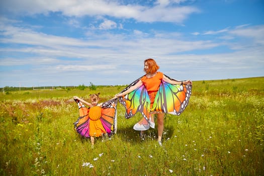 Happy female family with red haired mother and daughter with bright butterfly wings having fun on green and yellow meadow full of grass and flowers in sunny summer day. Concept family love