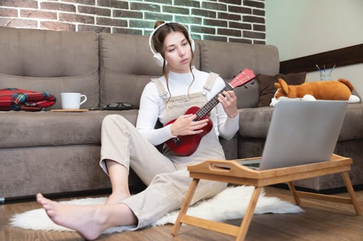 Hobbies and leisure activities during quarantine. Online training, online classes. A young woman watches a video lesson on playing the guitar, she sits on a cozy plaid with a guitar.