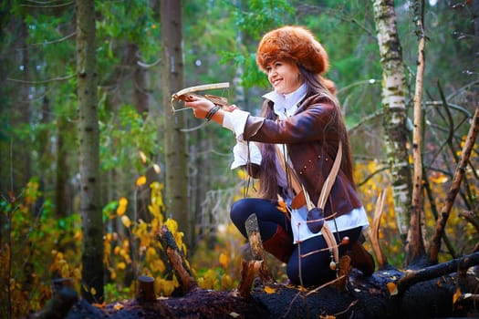 Girl in a leather jacket, a big red fox fur hat and with a crossbow in the forest in autumn. A female model poses as a fabulous royal huntress on nature hunt at a photo shoot