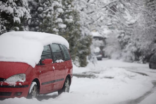 A red passenger car is visible in the foreground. The roof of the car is completely covered with a layer of snow. Blurred background. In the background you can see a snowy street and flakes of falling snow.