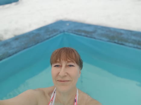 Adult mature woman takes selfie in a pool with warm hot termal mineral water in winter and white snow around. Wellness center and the concept of health care. Travel, recreation, medicine and rest