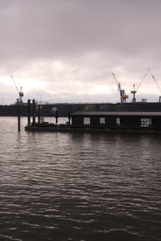 Vertical monochrome of a floating cafe on the water before the cranes in the port of Hamburg