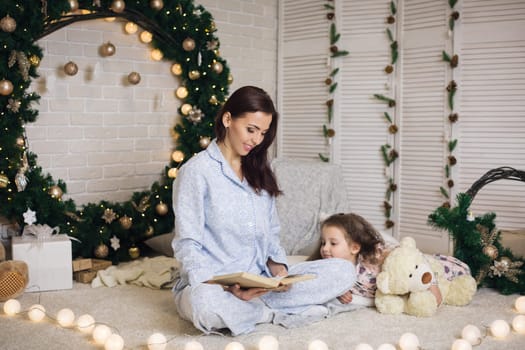 mother reading book to her little cute daughter near Christmas tree at home