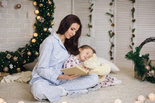 mother reading book to her little cute daughter near Christmas tree at home