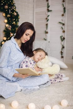 mother reading book to her little cute daughter near Christmas tree at home