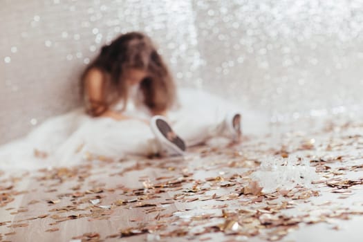blur sad little child girl in dress sitting on the floor with confetti on background with silver bokeh. focus on the crown in the foreground