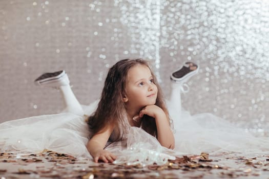 little girl in white dress lies on the floor and looks away on background with silver bokeh.